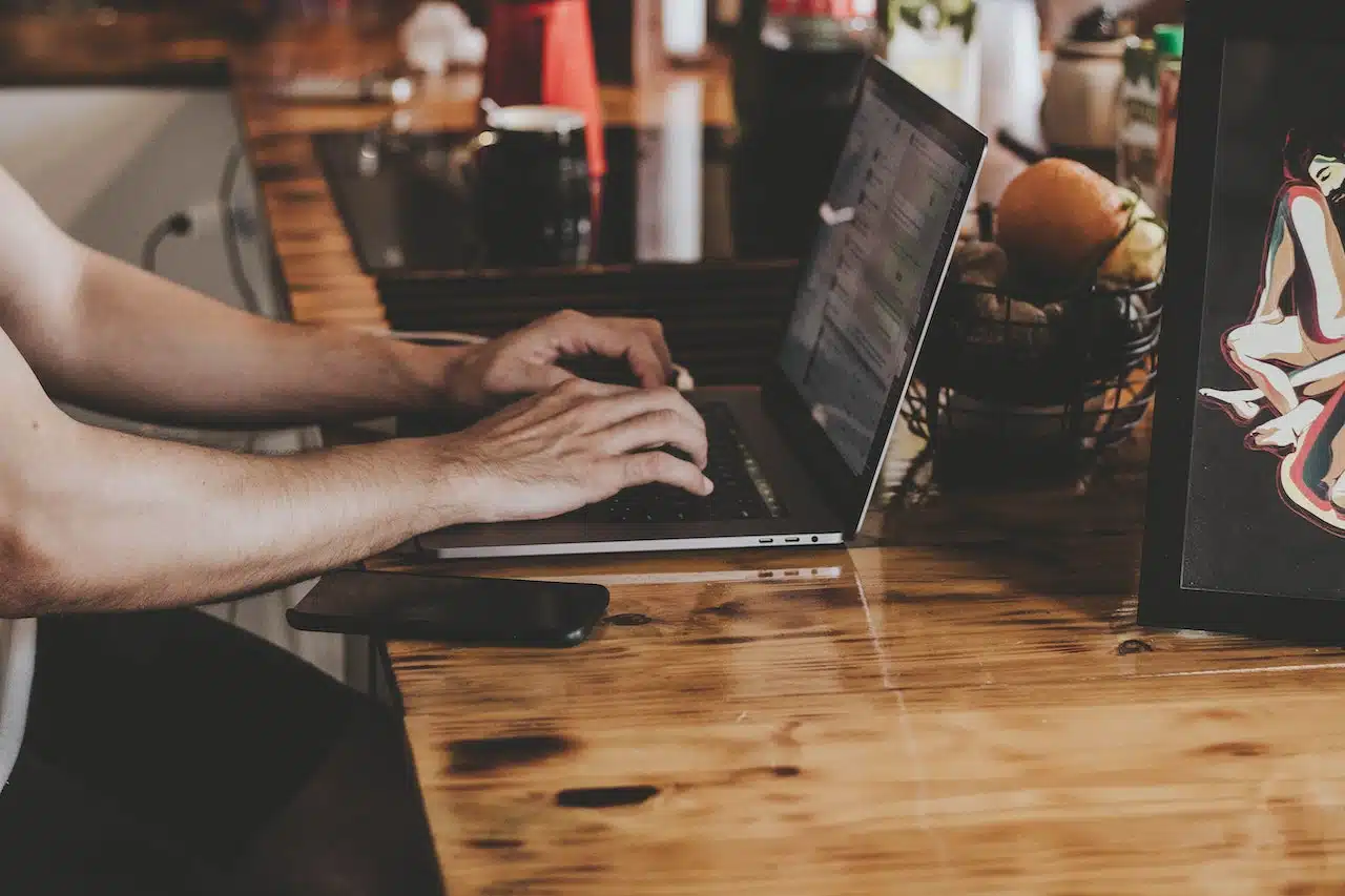 A person typing on a laptop at a wooden kitchen counter explores the possibilities of graph-as-a-service, with a smartphone nearby. A fruit basket adds color, while a picture frame with a vibrant image hangs to the right.
