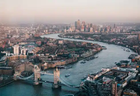 Aerial view of London with the River Thames winding through the city. The iconic Tower Bridge is visible in the foreground. The skyline includes various buildings and structures, with a hazy sky above.