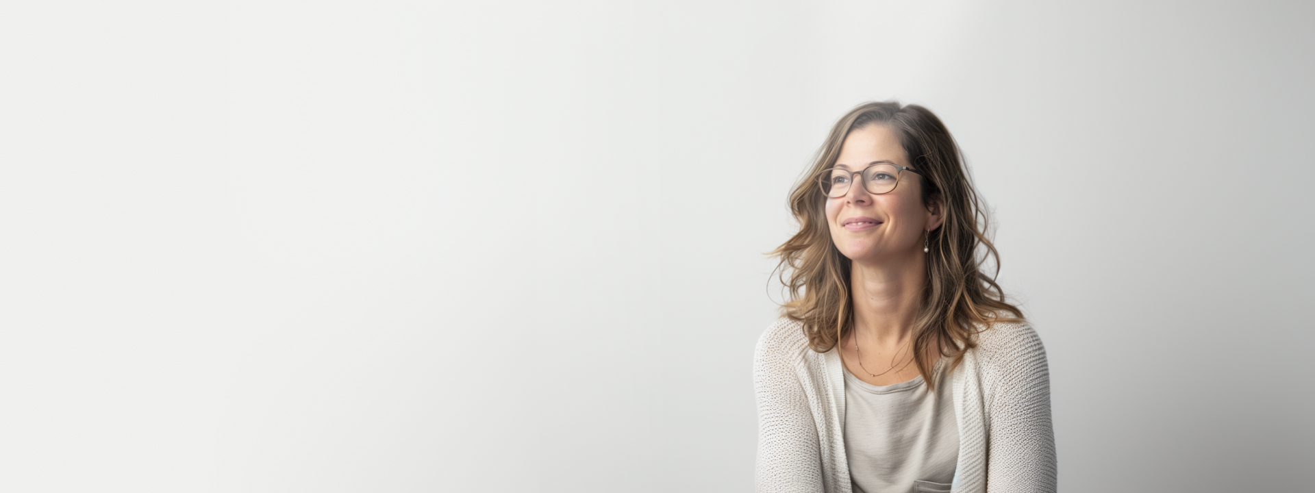 A woman with glasses and wavy hair sits smiling against a plain, light background. She is wearing a light-colored sweater over a shirt. The photo is bright, with ample negative space on the left side.