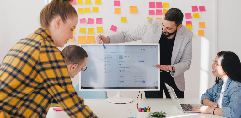 Four colleagues collaborate in an office with colorful sticky notes on the wall. One person presents at a screen, while the others engage at the table. The atmosphere appears positive and cooperative.